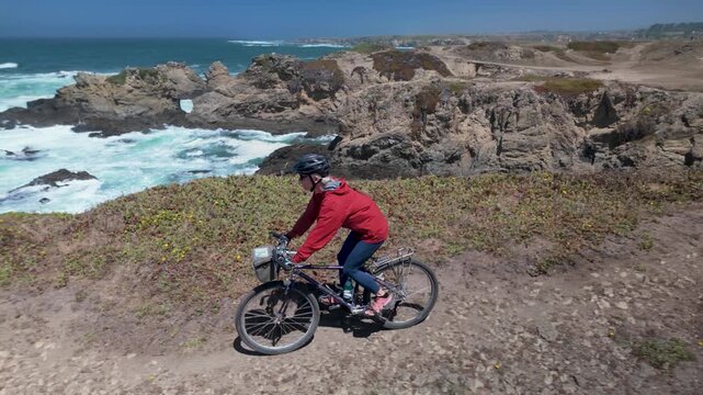 A woman rides her bike along the cliffs in Fort Bragg. The ocean waves crash below, creating a lively outdoor adventure.