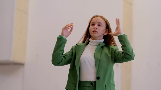 Young student in green jacket stretches her long hair to the sides and looks at herself with a serious expression, softly singing along to music in her head and imagining a stage or runway role.