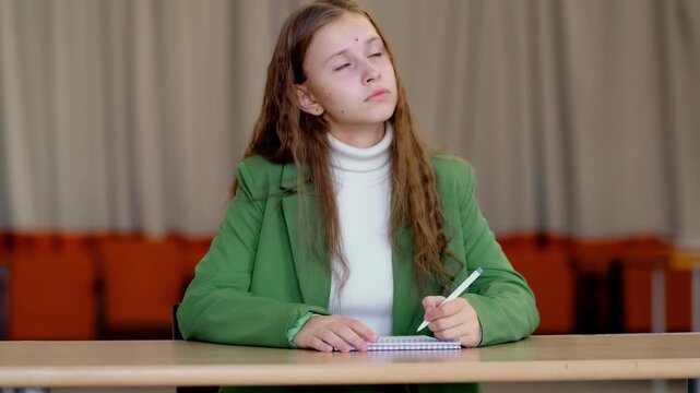 Young girl in green jacket sits in empty classroom with notebook, looks up with tired bored expression, theme lack of motivation and burnout from endless school assignments.