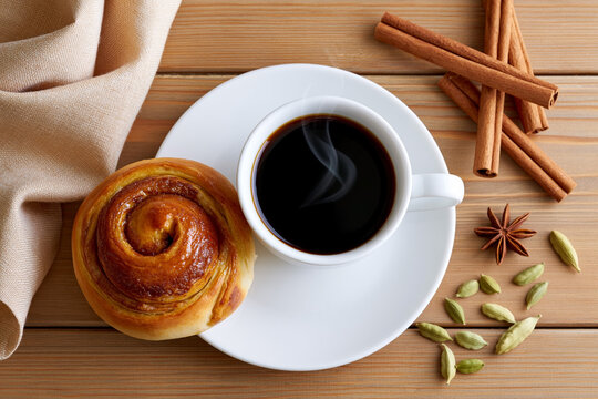 Freshly brewed coffee in a white cup beside a cinnamon roll on a white plate, surrounded by spices including cardamom and cinnamon sticks on a wooden table