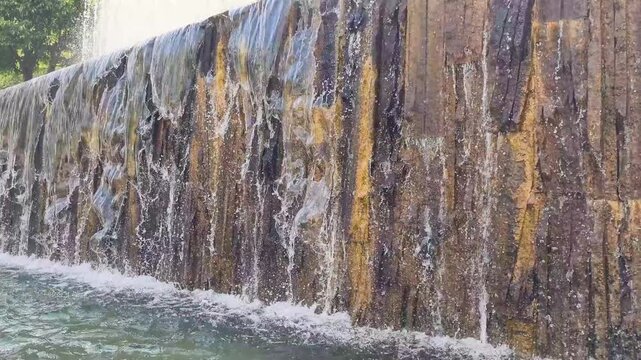 Water flowing down a textured stone wall creating a refreshing artificial waterfall in a modern city park fountain, creating a tranquil and relaxing atmosphere