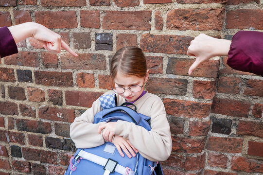 A lonely teenage schoolgirl in glasses stands by a wall outdoors while a group of classmates point, laugh, and mock her, conveying bullying, social isolation, and emotional distress.