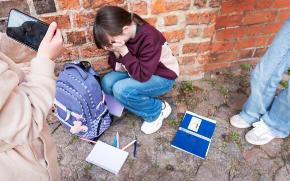A lonely teenage schoolgirl sits by a wall outdoors while a group of classmates point, laugh, make video and mock her, conveying bullying, social isolation, and emotional distress.