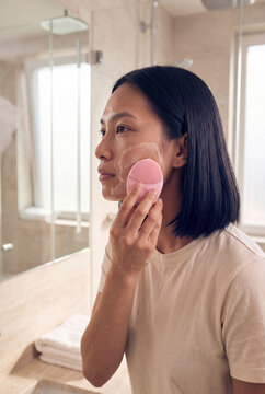 Asian woman applying facial cleanser with pink sponge in bathroom mirror for skincare routine poster and personal care banner
