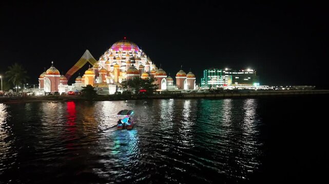 Aerial view of the illuminated Masjid Kubah 99 Asmaul Husna mosque at night with a small boat on the water in Kecamatan Mariso, Sulawesi Selatan, Indonesia.