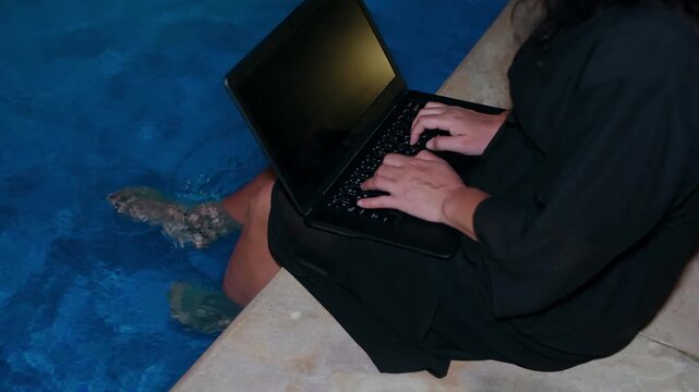 Man works on laptop poolside at night, feet in illuminated blue water, wearing dark clothing against textured concrete edge.