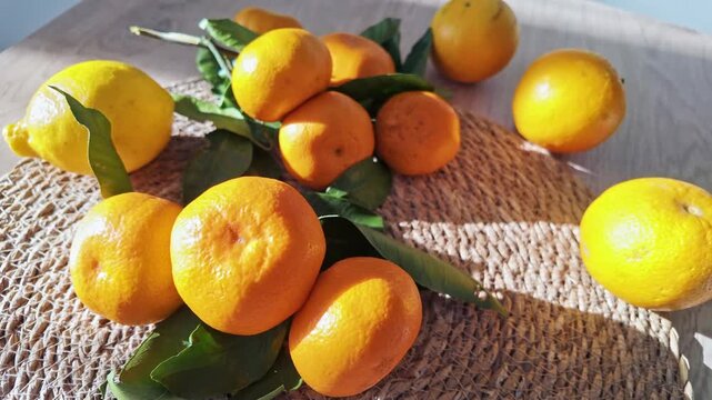 Zoom into sunlit lemons and mandarins with green leaves resting on woven straw mat.