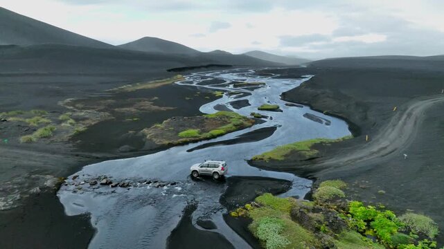 Aerial view of a 4x4 vehicle crossing a shallow river in a vast volcanic landscape with black sand and patches of green moss in Hella, Rangarthing ytra, Iceland.
