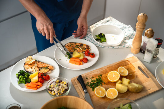 Healthy Eating with a Caregiver Arranging Grilled Salmon, Broccoli, Carrots, Potatoes and Roasted Tomatoes on Plates in a Bright Home Kitchen