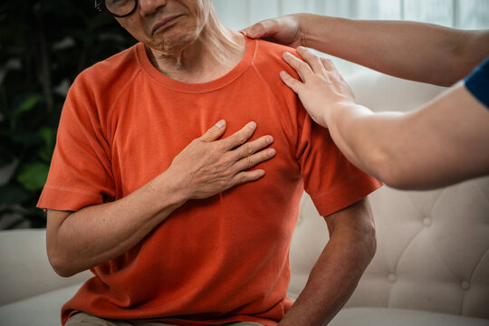 Senior Healthcare Support for an Asian Elderly Man Holding His Chest while a Caregiver Places Both Hands on His Shoulder during a Home Visit Indoors