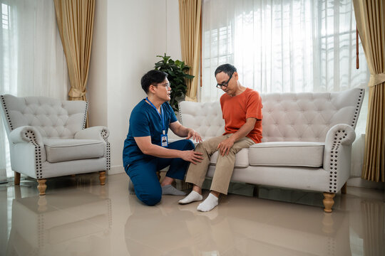 Male Nurse Assessing the Knee of an Asian Elderly Man during a Home Healthcare Visit while the Patient Sits on a Sofa in a Spacious Living Room