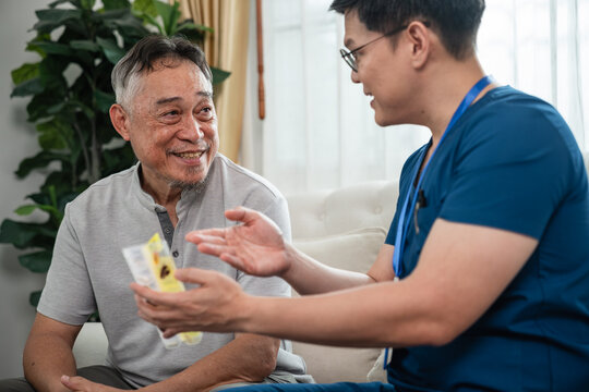 Male Nurse Explaining Medication to an Asian Senior Man during a Home Healthcare Consultation in a Bright Living Room with Medicine Packaging in Hand