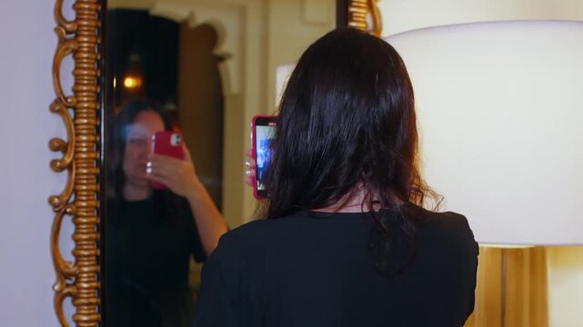 Person wearing black clothing holds smartphone up to ornate golden mirror, capturing self portrait reflection in interior space with warm lighting.