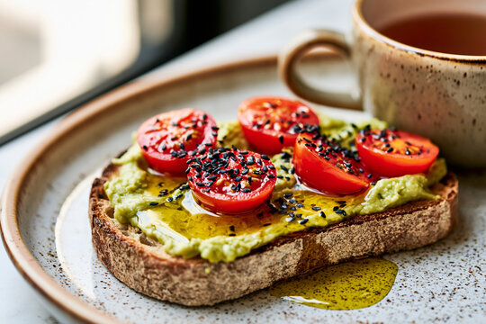 A slice of artisan bread topped with mashed avocado, ripe cherry tomatoes, a sprinkle of black sesame seeds and olive oil.