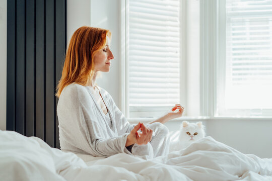 Middle aged woman in pajama and her white cat pet meditating on bed, practicing mindfulness and relaxation for a peaceful morning self-care and wellness routine at home. Mental health practice routine