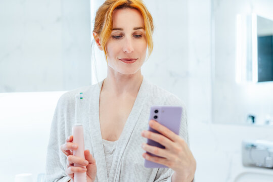 Middle aged woman puts settings of electronic toothbrush with mobile phone app. Wireless connecting sonic toothbrush with smart phone app. Modern home health care technology concept. Selective focus