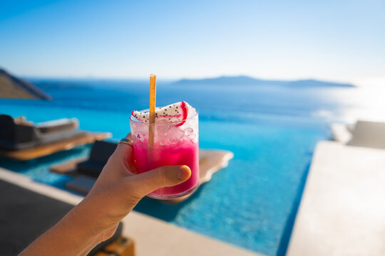 A woman's hand holds a fresh cocktail with ice. Pool with blue water and sea view in the blurred background. Santorini island, Greece.