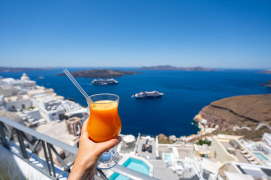Santorini island, Greece. Woman's hand holds a glass of fresh orange juice. White architecture and the blue sea with sky in the blurred background.