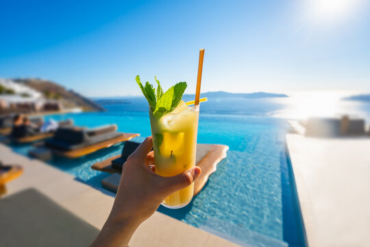 Woman's hand holds a fresh cocktail with ice. Pool with blue water and sea view in the blurred background. Santorini island, Greece.