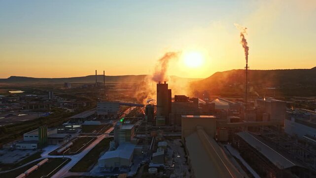 Smoke rises from tall chimneys as machinery operates in a factory setting during sunset. The sun shines brightly behind the mountains and industrial buildings.