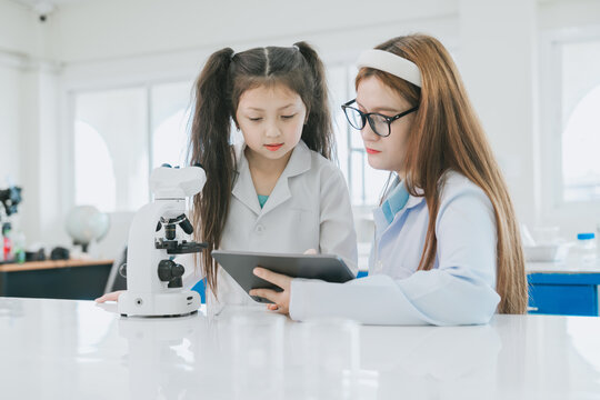 Curious young student in white coat uses tablet computer beside microscope in science laboratory classroom during school lesson