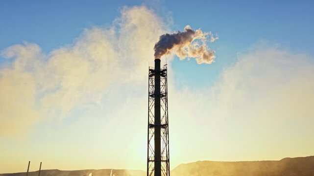 Smoke is coming out of a tall chimney at a factory as the sun sets. The sky is clear with some clouds creating a golden hue. The landscape has hills in the background.