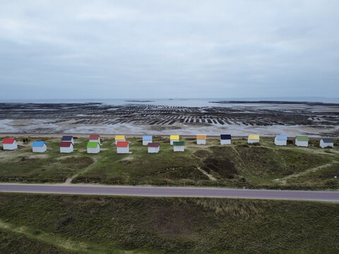 Aerial view of the iconic colorful beach huts lined up along the grassy dunes with oyster beds visible at low tide in Gouville-sur-Mer, Normandy, France.