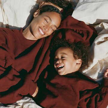 Smiling mother and child lying together in burgundy sweaters on a bed in warm sunlight for family lifestyle background and cozy banner