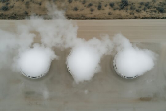 Aerial view of three steam vents releasing white vapor over an industrial site, with a barren landscape in the background.