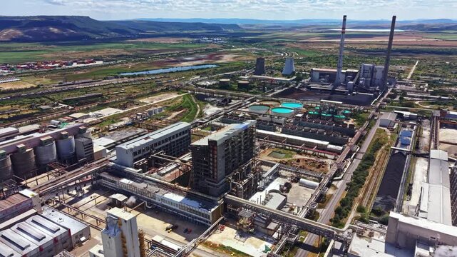 Workers and machines operate in a big industrial zone surrounded by fields and hills under a blue sky with some clouds. The site includes various structures and processes in action.