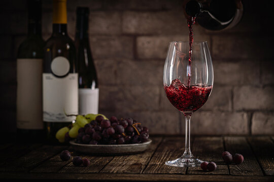 A dark, elegant still life of red wine being poured into a glass with wine bottles and fresh grapes in the background. Perfect for wine-themed compositions