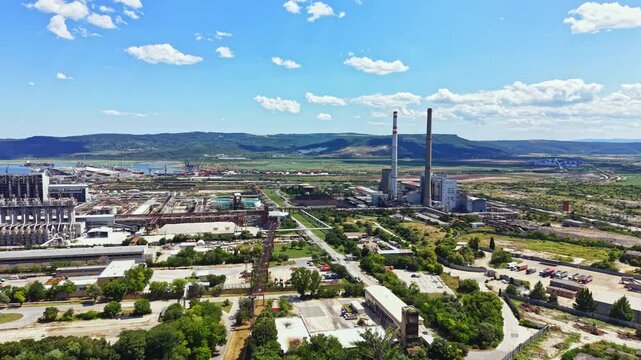 View shows an industrial zone near the coast with various factories and a power plant. The scene captures clear skies and green land between structures.