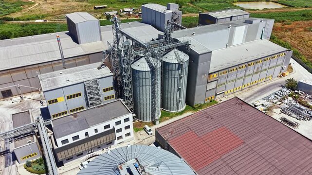 An industrial area with silos and large structures. Machinery and equipment are visible in the background. Green fields can be seen beyond the buildings.