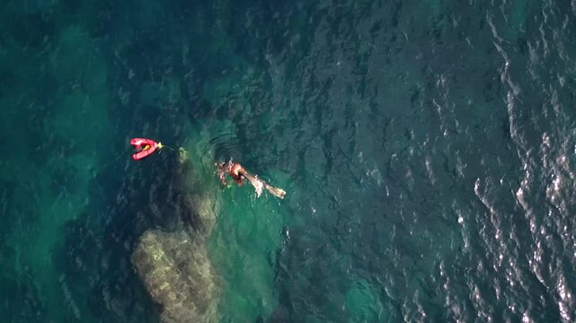 Aerial view of a person snorkeling with fins and a red safety buoy in the clear blue waters of Cala Llebeig in El Poble Nou de Benitatxell, Valencian Community, Spain.