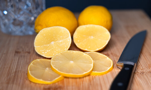 Close-up of two lemons a kitchen knife on a cutting board, with sliced lemon wedges in the foreground. Drinking glass in the background. Subtropical fruit, vitamin C, freshness, citrus fruit, healthy.