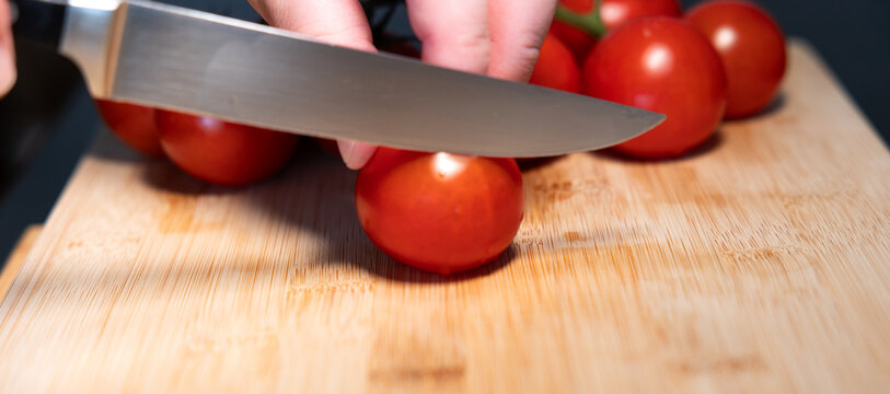A hand is holding a red tomato on a cutting board and begins to slice the tomato with a kitchen knife. Freshness, vegetable, contains vitamin C, potassium. Healthy eating, healthy lifestyle, raw food.