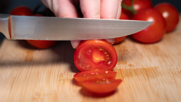 A hand is holding a red tomato on a cutting board and slicing it with a kitchen knife. Freshness, vegetable, contains vitamin C, potassium, vegetarian food, healthy eating, aw food.
