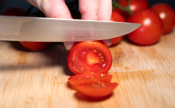 A hand is holding a red tomato on a cutting board and slicing it with a kitchen knife. Freshness, vegetable, contains vitamin C, potassium, vegetarian food, healthy eating, aw food.