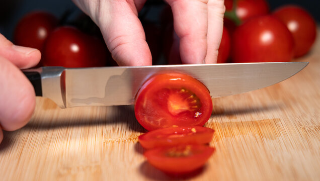 A hand is holding a red tomato on a cutting board and slicing it with a kitchen knife. Freshness, vegetable, contains vitamin C, potassium, vegetarian food, healthy eating, aw food.
