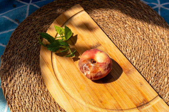 Ripe peach, Prunus persica, with a large soft brown lesion and decaying flesh caused by advanced fungal spoilage. The damaged fruit rests on a bamboo board with fresh mint, illustrating brown rot, foo