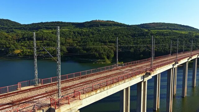 A railway bridge spans over a river, with steel beams and gravel on the tracks. Green hills rise on either side, showing trees under a clear sky.