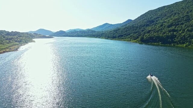 A boat travels on a river that stretches between hills. The water reflects sunlight while trees and land outline the riverbanks under a clear sky.