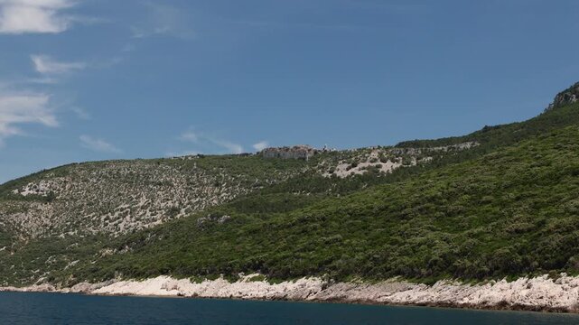 view of Lubenice historic village perched on cliffs from Sveti Ivan Beach. Providing a panoramic view of the rugged coastline. Lush green slopes. And the clear blue Adriatic Sea on Cres Island