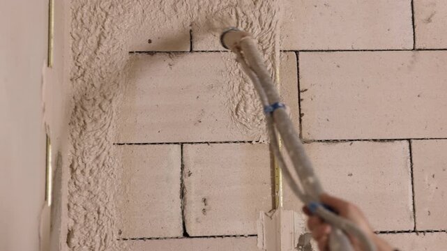 sprays putty, Concrete block, Interior renovation. Close-up spray hose coating indoor cinder block wall with wet textured plaster during renovation.