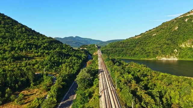 Train tracks stretch into the distance between hills covered in trees near a river on a bright sunny day in a rural landscape. This scene shows nature alongside human development.