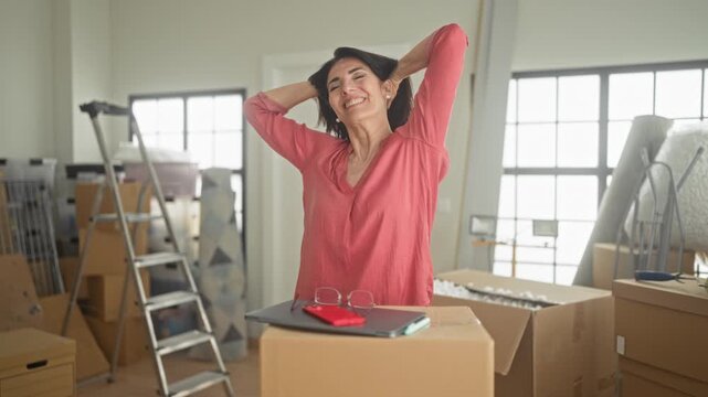 Woman stretching arms over a packed box, smiling amid stacked boxes, ladder and mattress in a building; new home relief.