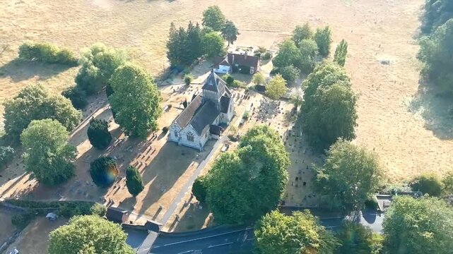 Aerial drone view of a historic village church and surrounding graveyard, framed by mature trees, countryside fields, nearby houses and rural roads on a sunny day