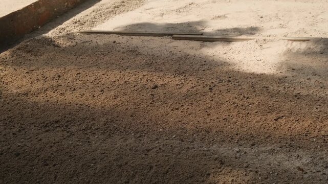 Portions of loose building sand fall onto the prepared base before paving stone installation. Leveling layer for sidewalk construction and municipal infrastructure.