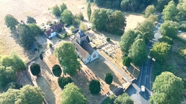 Aerial drone view of a historic village church and surrounding graveyard, framed by mature trees, countryside fields, nearby houses and rural roads on a sunny day