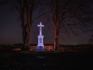 Przydrożna kapliczka z krzyżem (Crux) nocą pod gwieździstym niebem, ujęta pomiędzy starymi drzewami. Symbol wiary, tradycji i wiejskiego krajobrazu Polski. © Henryk Niestrój